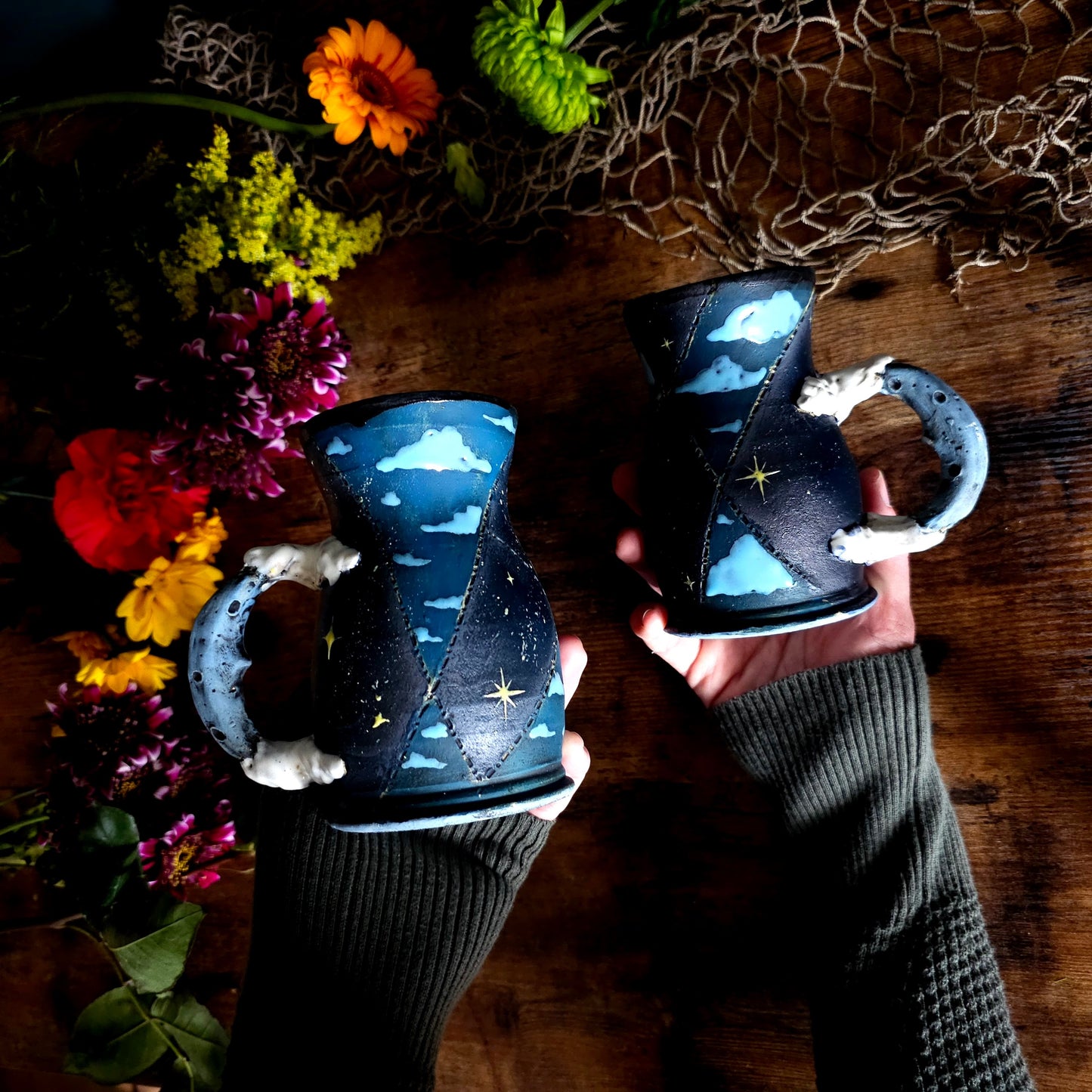 Handcrafted ceramic mug with a dark blue and black hourglass shape, star and cloud design, moon-inspired handle, styled on a rustic wood table with wildflowers