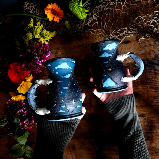 Handcrafted ceramic mug with a dark blue and black hourglass shape, star and cloud design, moon-inspired handle, styled on a rustic wood table with wildflowers