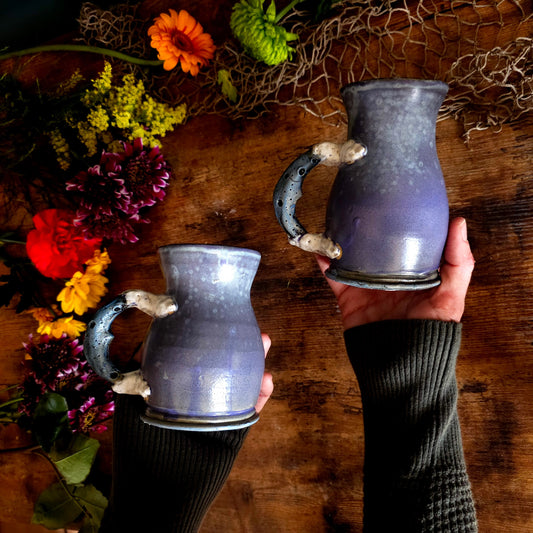 Handmade lavender ceramic mug with moon crater handle on rustic wood table, moody cottagecore backdrop with dark green wall and floral jar.